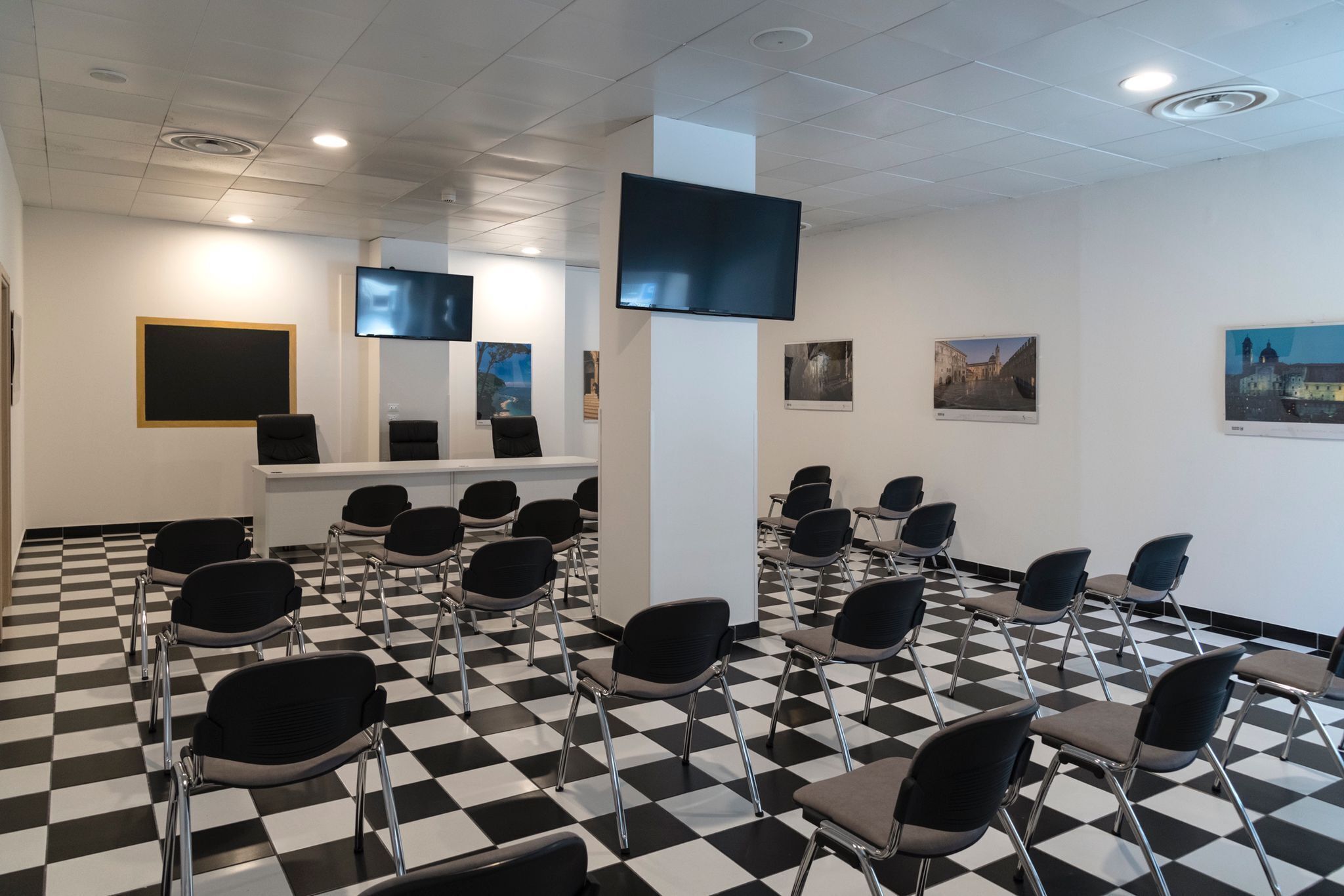 Una sala conferenze con pavimento a scacchi, file di sedie, un banco reception e un televisore a schermo piatto montato a parete.