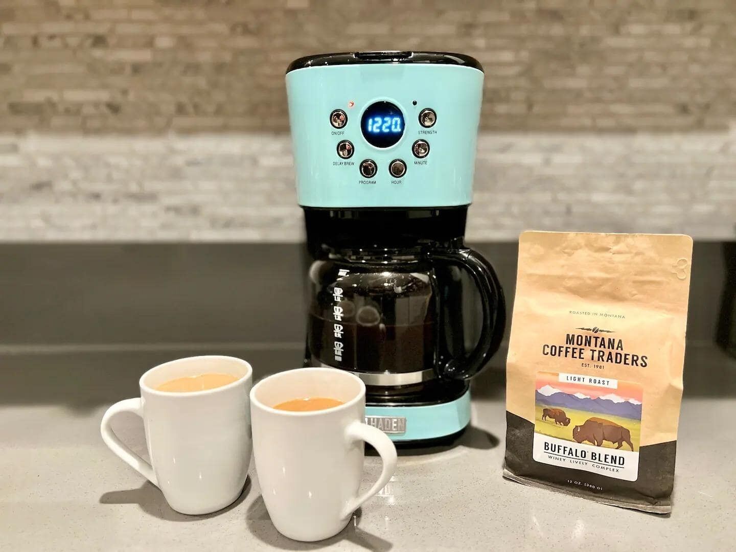 A coffee maker with a digital display, two white cups, and a bag of coffee labeled 'Montana Coffee Traders'.