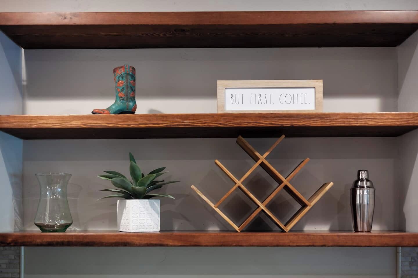 A shelf with a sign that reads "BUT FIRST COFFEE," a decorative plant, a wooden wine rack, and a cocktail shaker.