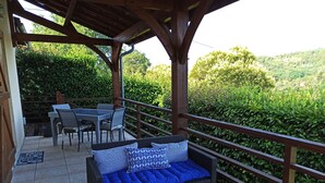 Een overdekt terras met een tafel en stoelen, een rieten bank met blauwe kussens en een balustrade met uitzicht op een weelderig groen landschap.