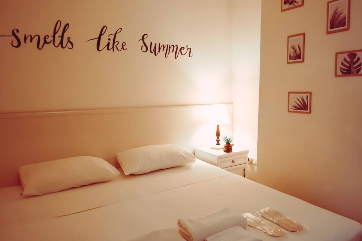 A neatly made bed with pillows, a bedside table with a lamp and a small plant, and a wall with a 'smells like summer' sign and leaf art.