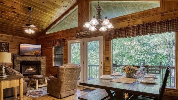 L’intérieur chaleureux d’un chalet avec une cheminée, des meubles en bois, une table à manger dressée pour quatre personnes et une grande fenêtre donnant sur un paysage verdoyant.