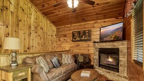 A cozy cabin interior with a stone fireplace, wooden walls, a leather sofa, and a television mounted above it.