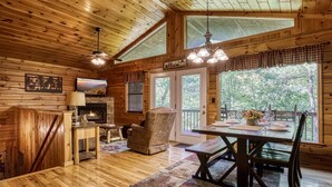 L’intérieur chaleureux d’un chalet, avec un plancher en bois, une cheminée, une table à manger entourée de chaises et de grandes fenêtres donnant sur le paysage extérieur.