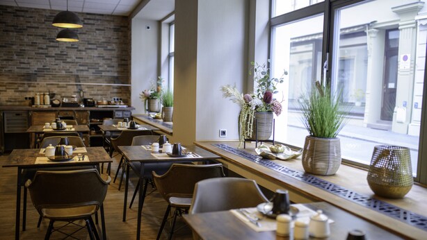 A modern cafe with wooden tables and chairs, a brick wall, and large windows offering a view of the street. - Stadthotel König Albert (Zittau)