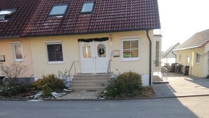 Une maison à deux étages, avec un toit recouvert de tuiles rouges, un porche à l’avant et un petit jardin.