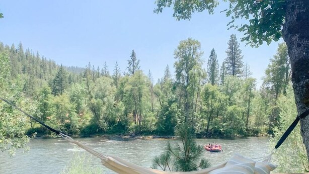 Un hamac est tendu entre deux arbres, offrant une vue sur une rivière, et on aperçoit un radeau au loin.