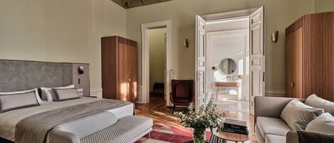 A spacious bedroom with a large bed, a sofa, a small table with a vase, and a bathroom visible through an open door.