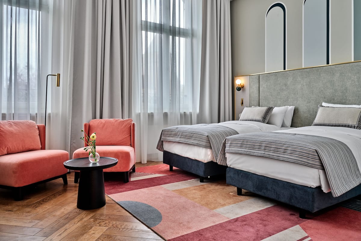 A modern hotel room with a large bed, a seating area with a chair, a small table, and a large window with curtains.