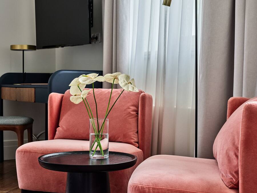 A modern living room with a pink sofa, a black coffee table, and a glass vase with white flowers. A flat-screen TV is mounted on the wall, and a floor lamp is visible.