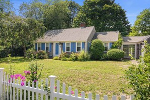 A two-story house with a front porch, surrounded by a white picket fence and various shrubs.