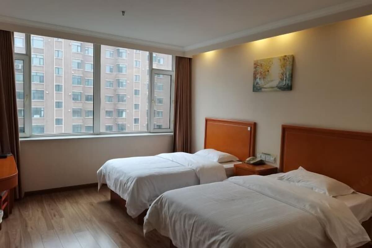 A hotel room with two beds, a window with curtains, a painting on the wall, and a wooden floor.