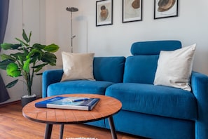 Un canapé bleu avec des coussins blancs, une table basse ronde en bois sur laquelle repose un livre, une plante en pot et des œuvres d’art encadrées accrochées au mur.
