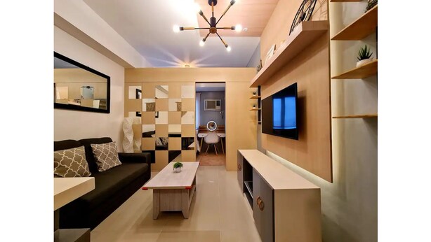 Ein modernes Wohnzimmer mit Sofa, Couchtisch und Fernseher. Es gibt einen Essbereich mit einem Stuhl und einem kleinen Tisch. Die Wohnung verfügt über eine Deckenleuchte und Regale mit Dekoration.