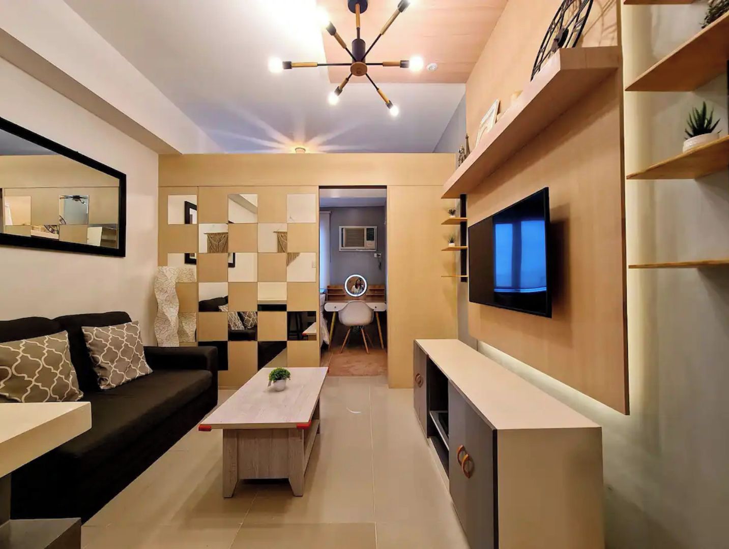 A modern living room with a sofa, coffee table, and TV. There is a dining area with a chair and a small table. The room has a ceiling light fixture and shelves with decorative items.