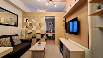 A modern living room with a sofa, coffee table, and TV. There is a dining area with a chair and a small table. The room has a ceiling light fixture and shelves with decorative items.