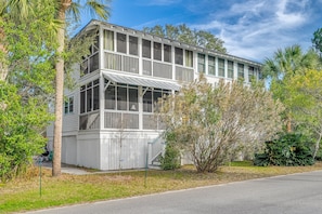 A two-story building with large windows and a balcony, surrounded by palm trees and other greenery.