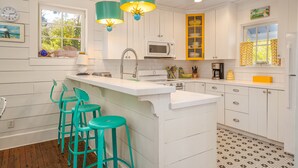 A kitchen with white cabinetry, a central island with bar stools, a microwave, and a window with curtains. - Private Pool + Whimsical Sleeping Porch Steps from Beach by Tybee Cottages (Tybee Island)