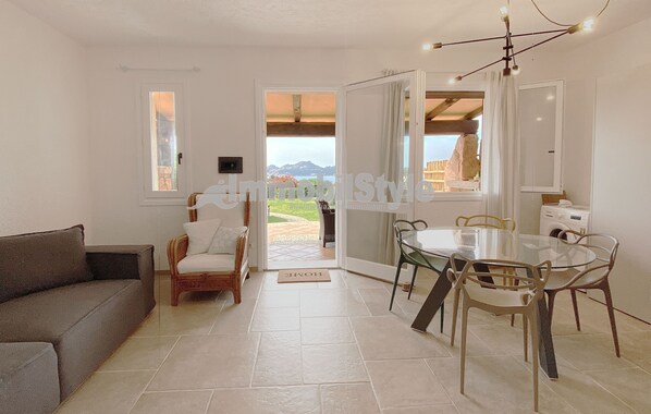 A modern living room with a sofa, wicker chairs, a glass dining table with metal chairs, a kitchen area with a white refrigerator, and a view of mountains through the open door.