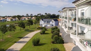 A multi-story hotel with balconies, a well-maintained lawn, and a view of surrounding houses. - Résidence Madame Vacances Les Appartements de Fontenelles (L'Aiguillon-sur-Vie)