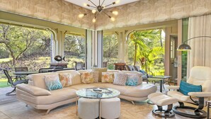 A spacious living room with a large beige sectional sofa, a glass coffee table, and a white armchair. There is a dining area with a table and chairs, and a view of a lush garden through large windows.