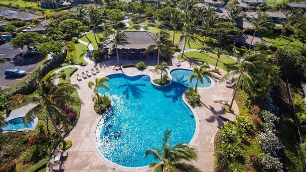 An aerial view of a resort with a central swimming pool, surrounded by palm trees and a golf course.