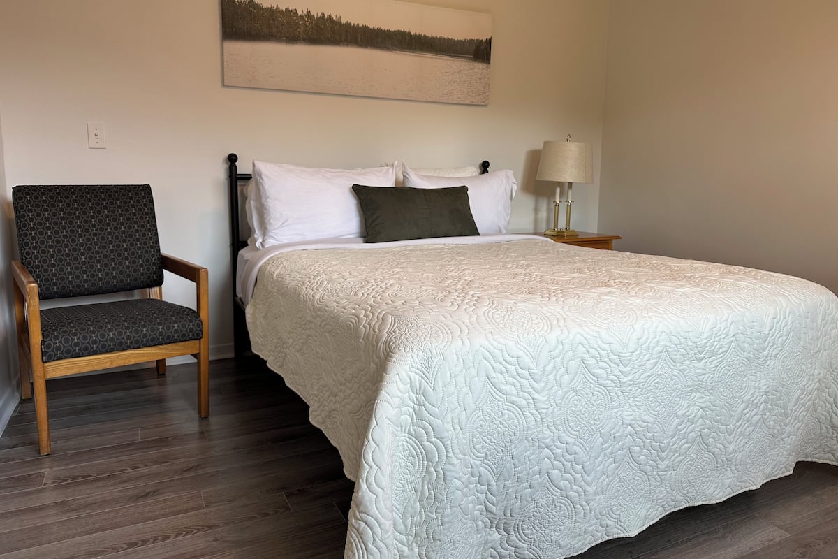 A bedroom with a large bed, a chair, a nightstand, a lamp, and a painting on the wall.