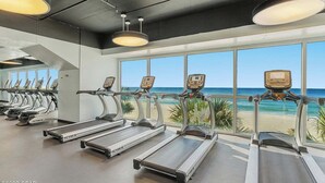 Un gimnasio con cintas de correr alineadas frente a una pared con ventanas amplias que dan a una playa.