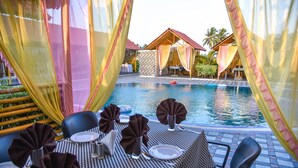 A poolside dining area with tables set for two, a pool, and a tropical setting with palm trees. - Pride Divine Resort Somnath (Veraval)