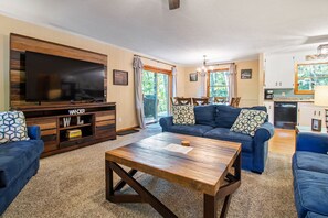 Una sala de estar con un televisor de pantalla plana grande, una mesa de centro de madera y un sofá azul con cojines estampados.