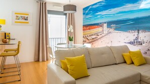 A living room with a beige sofa, yellow cushions, a glass table, and a chair. There is a large window with curtains, a framed picture on the wall, and a mural of a cityscape and beach. - Vita Portucale | Downtown Premium with Garage (Lisboa)