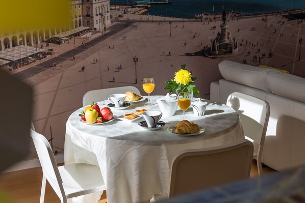 A table set with breakfast items, including fruits, pastries, and a glass of orange juice, overlooking a beach and historic buildings.