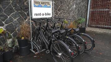 Letrero que dice 'Little Amsterdam' con un servicio de 'rent-a-bike', varias bicicletas y plantas en macetas, todo frente a un muro de piedra.