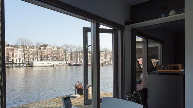 Une chambre moderne dotée d’une grande fenêtre donnant sur une rivière, d’une terrasse en bois et d’une vue sur le paysage urbain.