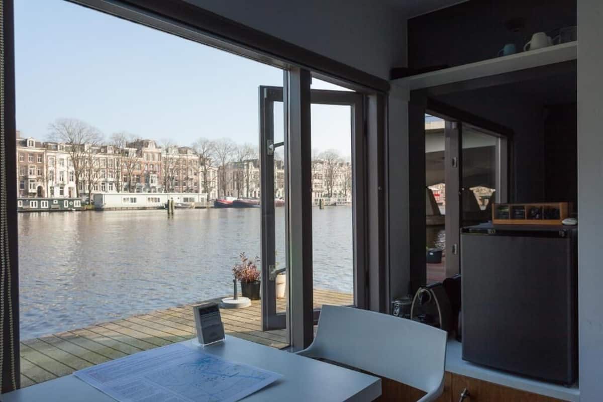 A modern room with a large window overlooking a river, a wooden deck, and a view of a cityscape.