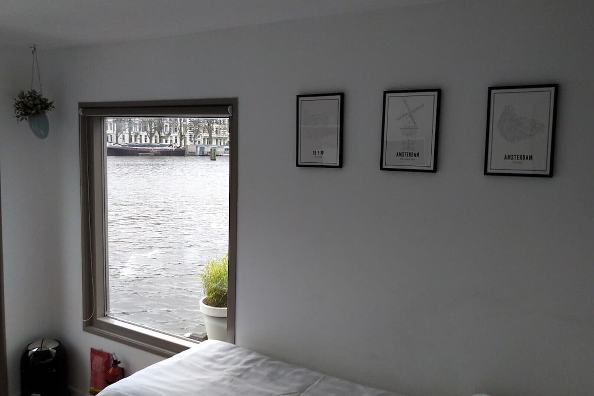 A room with a large window overlooking a canal, a bed, and three framed pictures on the wall.