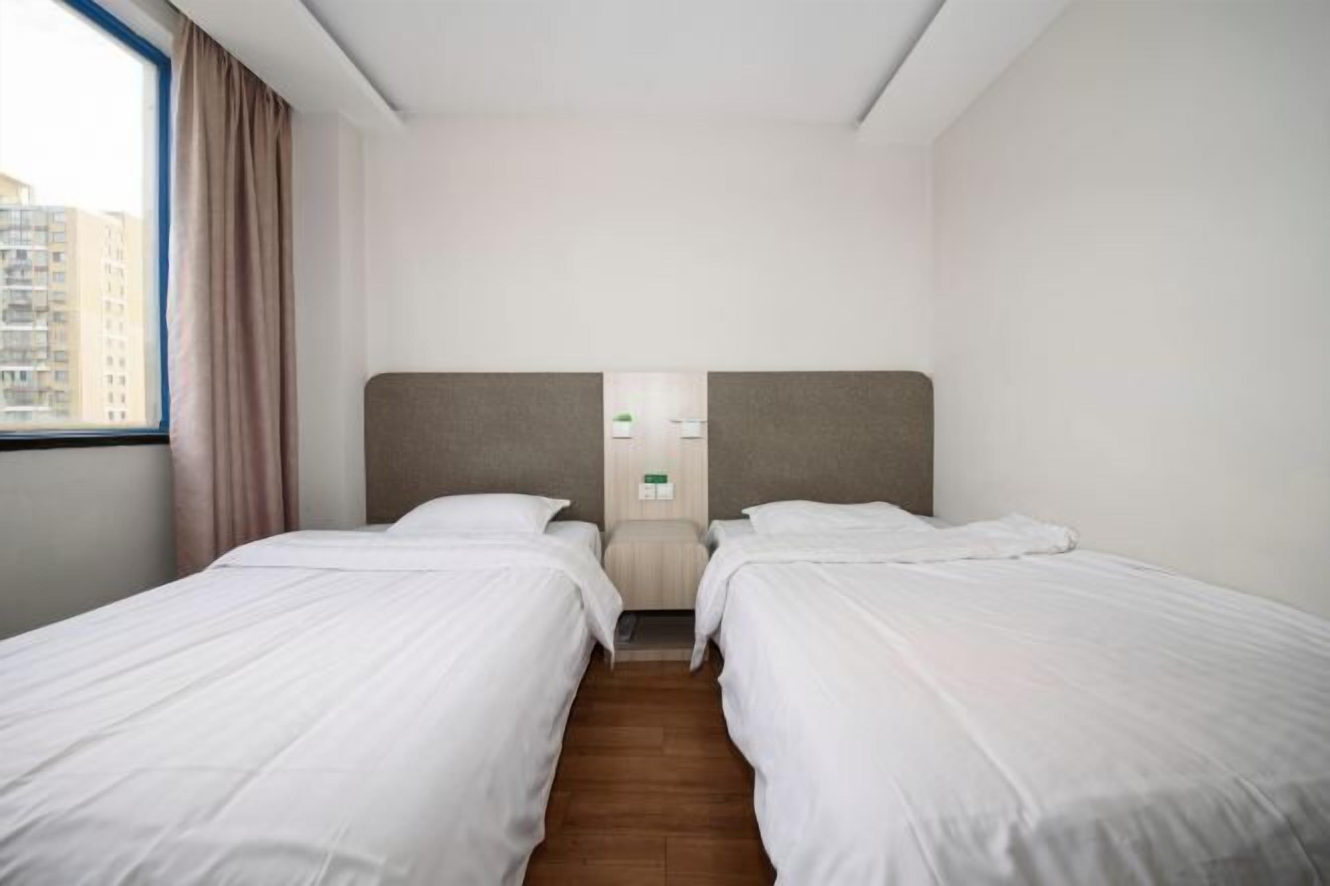 Habitación de hotel con dos camas individuales, piso de madera, ventana con cortinas y vistas a edificios.