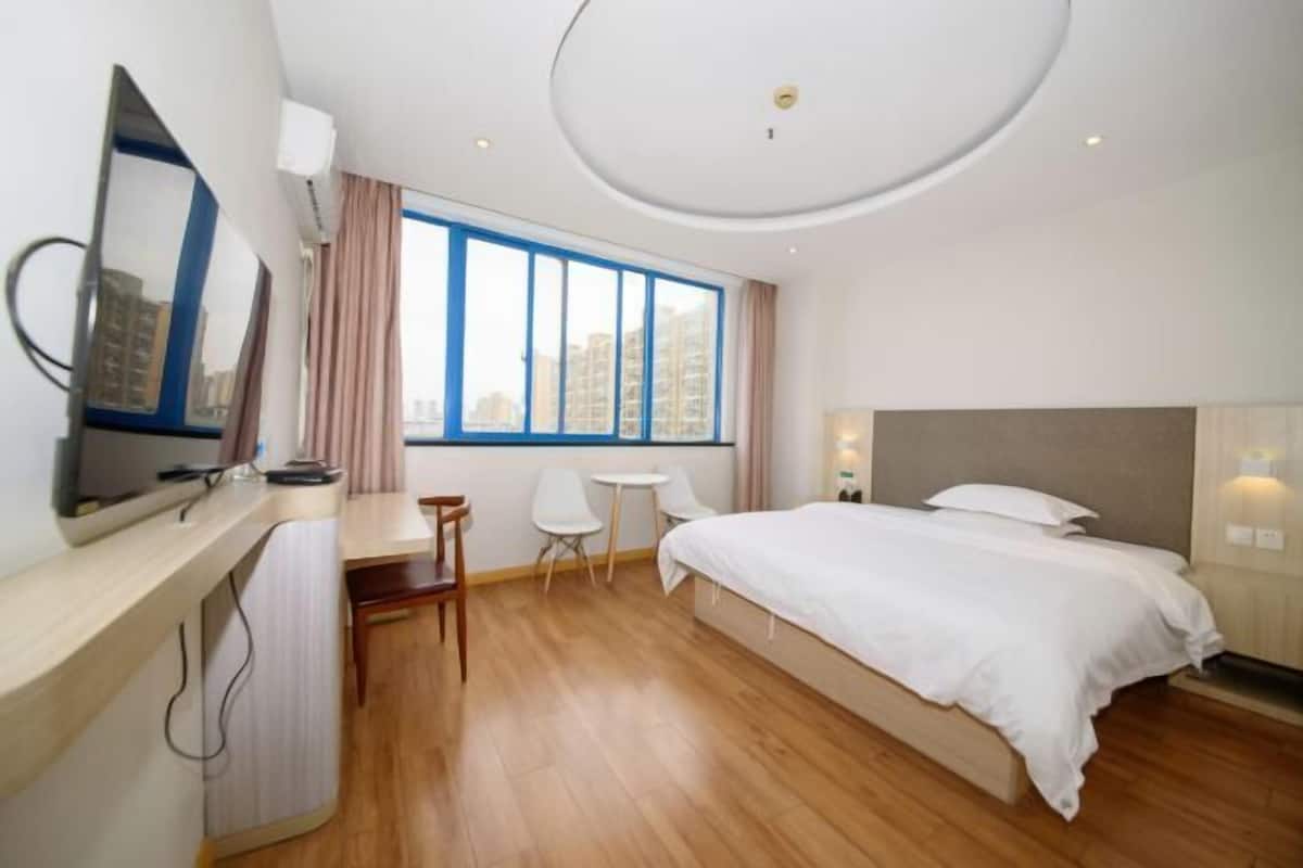 A modern hotel room with a large bed, a desk with a chair, a small table, and a large window offering a city view.