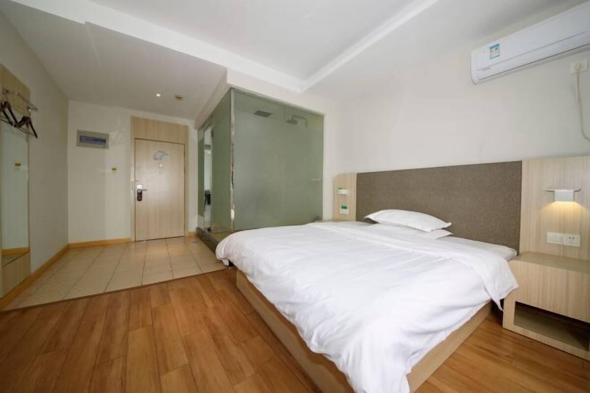 A hotel room with a large bed, wooden flooring, a glass door leading to a bathroom, and a wall-mounted air conditioner.