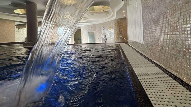 Una piscina interior con caída de agua, rodeada por un muro de azulejos e iluminación empotrada en el techo.