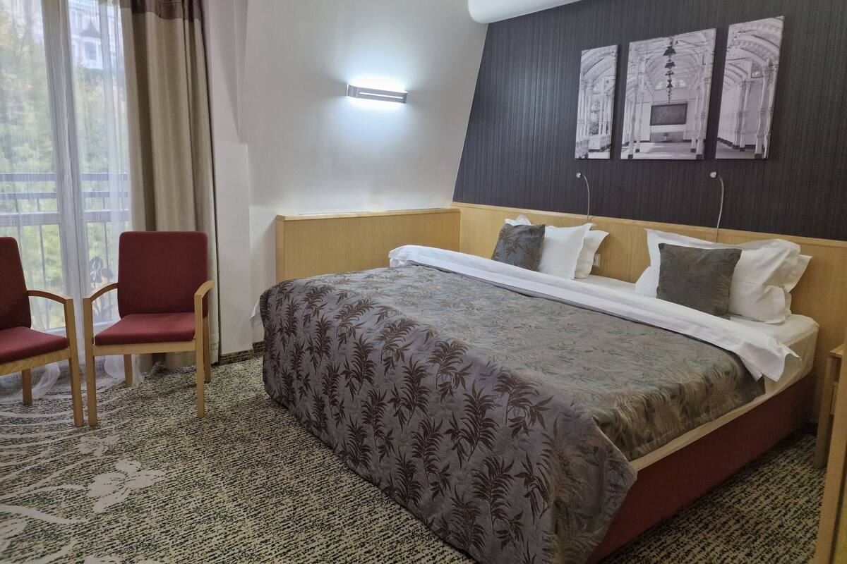 A hotel room with a bed, a chair, a wall-mounted light, and two framed pictures.