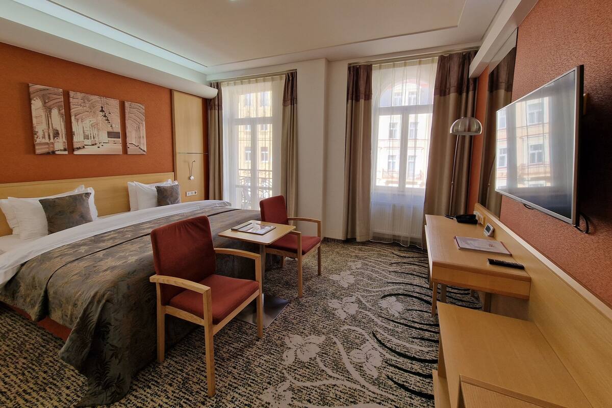 A hotel room with a bed, two chairs, a desk, a TV, and artwork on the wall.