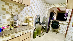 A compact kitchen with wooden cabinets, a gas stove, a refrigerator, and a small dining area with a chair. - Stay.Plus Kings & Queen's Apartment (Nairobi)