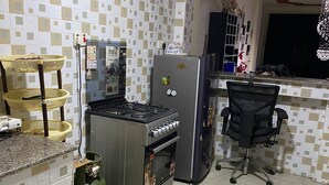 A kitchen with a gas stove, refrigerator, and a gas cylinder. There is a chair and a small table with a basket. - Stay.Plus Kings & Queen's Apartment (Nairobi)