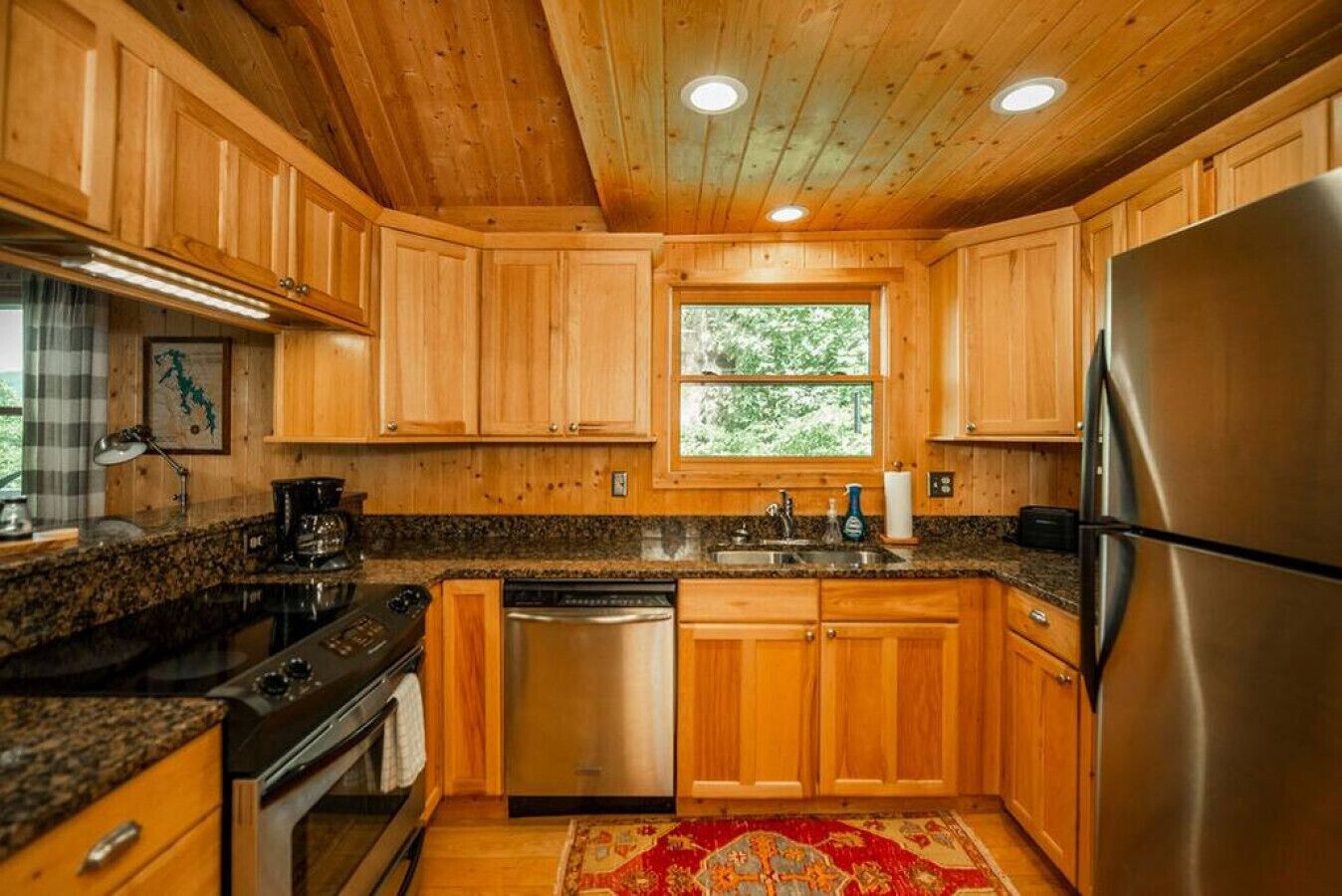 Cucina con mobili in legno, piani in granito, elettrodomestici in acciaio inossidabile e una finestra vista verde.