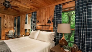 A wooden cabin bedroom with a bed, two bedside lamps, a chair, and a window with curtains. - Beautiful view with firepit and hot tub. Only 7 minutes to Cashiers and Glenville! (Cashiers)