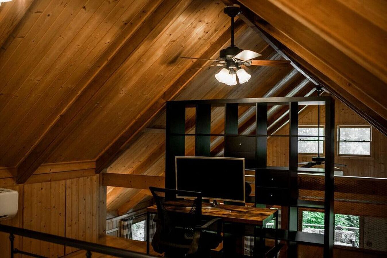 Un interior de madera con techo inclinado, un escritorio con computadora y una ventana que da a zonas verdes.