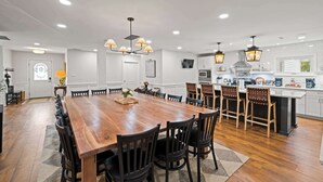 Un comedor amplio con una mesa de madera alargada rodeada de sillas, una cocina con armarios blancos y electrodomésticos modernos, e iluminación colgante.