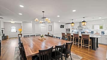 A spacious dining area with a long wooden table surrounded by chairs, a kitchen with white cabinets and modern appliances, and pendant lighting.