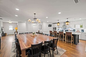 Un comedor amplio con una mesa de madera alargada rodeada de sillas, una cocina con armarios blancos y electrodomésticos modernos, e iluminación colgante.